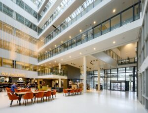 Moderne, lichte bedrijfs- of kantoorlobby aan de Thomas R. Malthusstraat met zitgedeelte, glazen gevel en een verlichte kunstinstallatie.